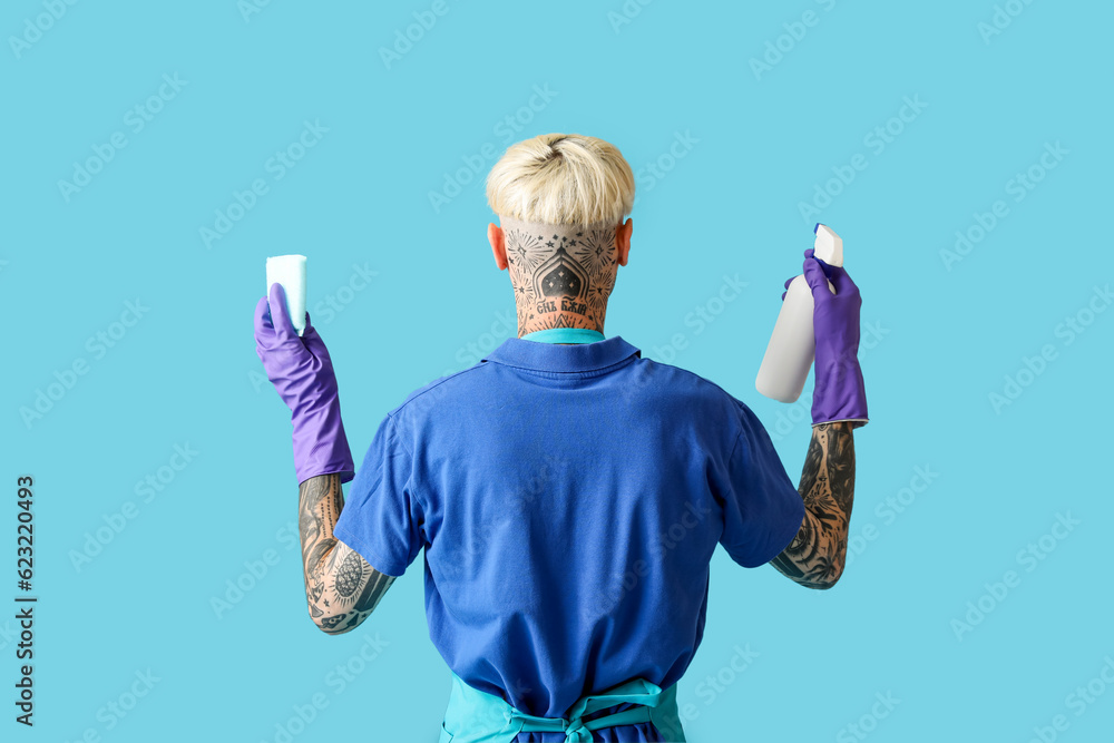 Young tattooed man with bottle of detergent and cleaning sponge on blue background, back view