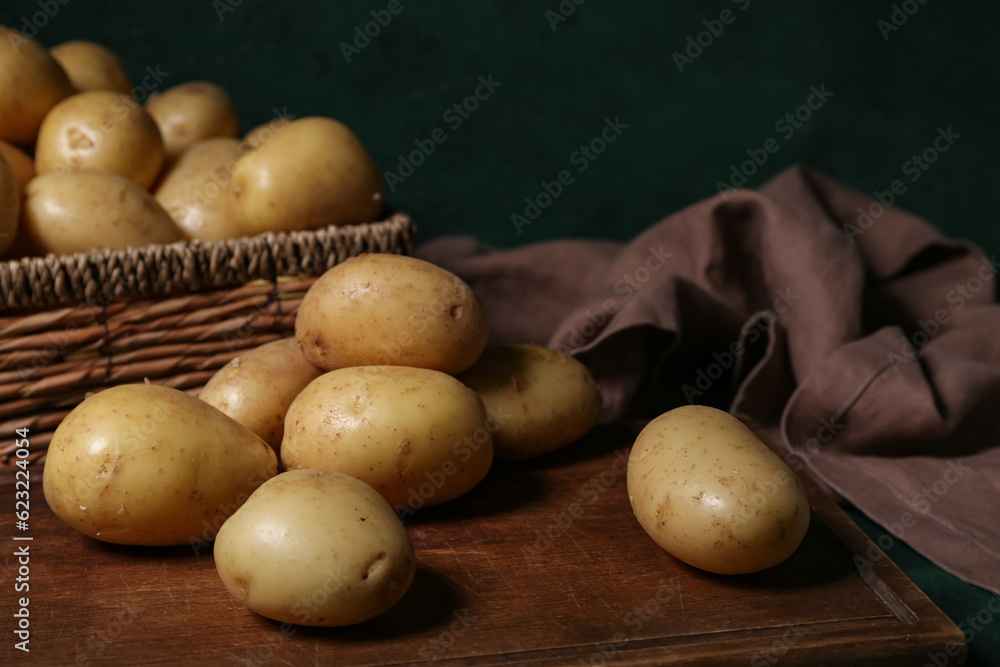 Wooden board and wicker box with raw potatoes on green background, closeup