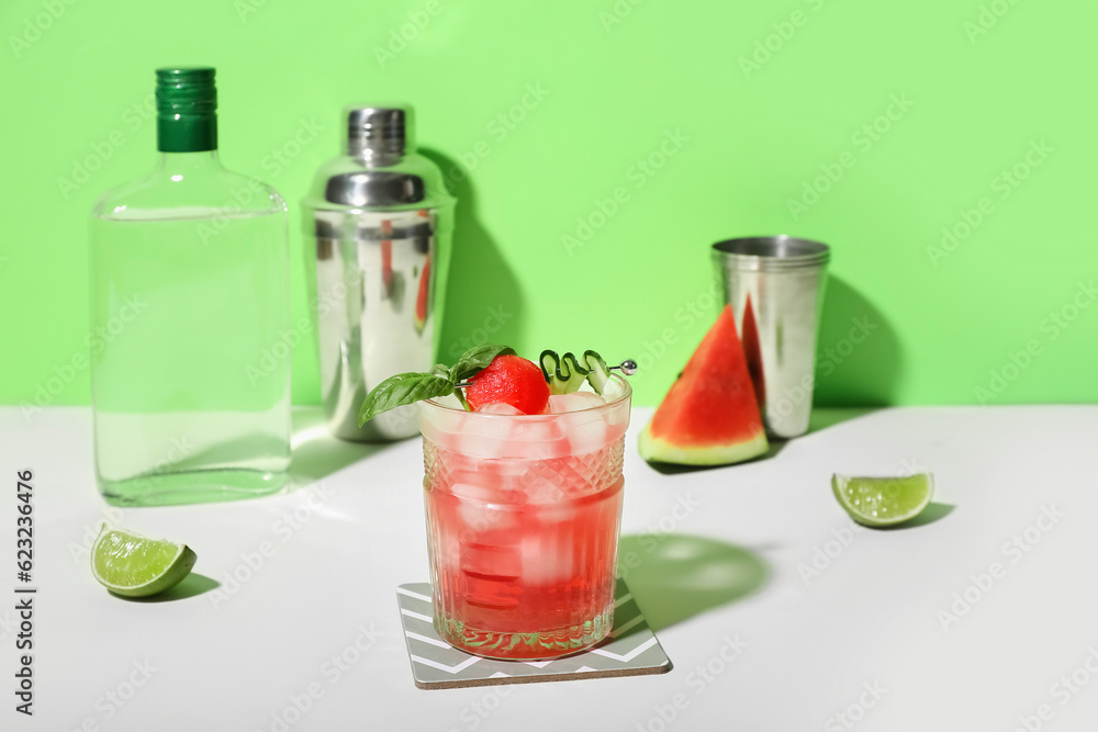 Glass of tasty watermelon cocktail with shaker and lime on white table near green wall