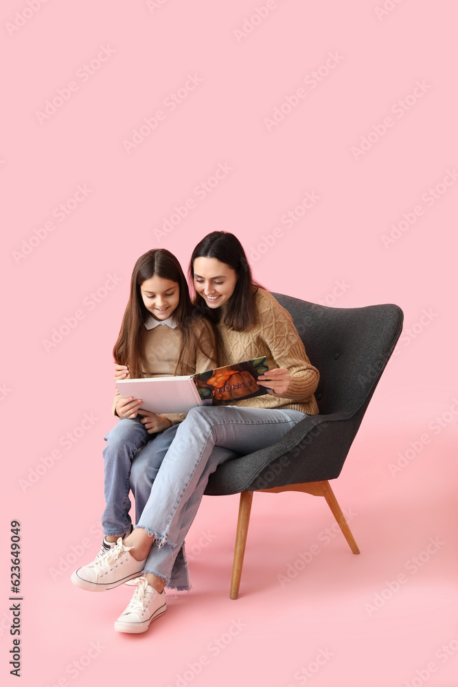 Little girl and her mother reading book in armchair on pink background