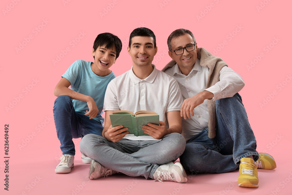 Happy little boy with his dad and grandfather reading book on pink background