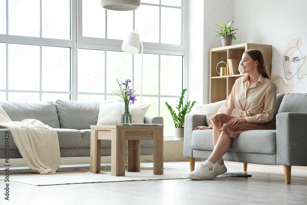 Happy young woman sitting on grey sofa in interior of light living room