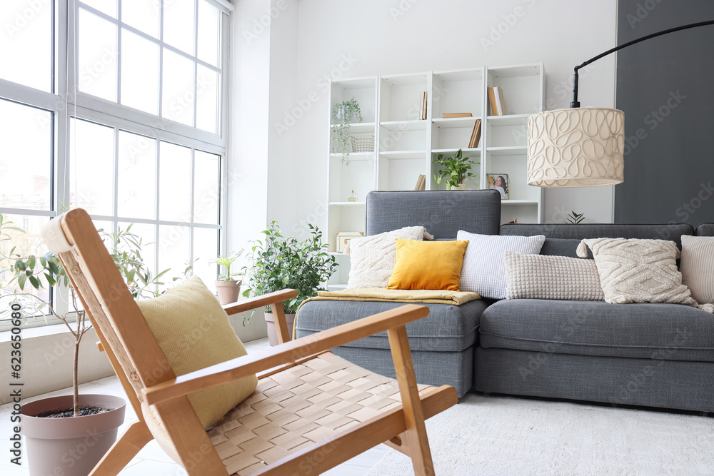 Interior of stylish living room with cozy grey sofa and wooden armchair near big window