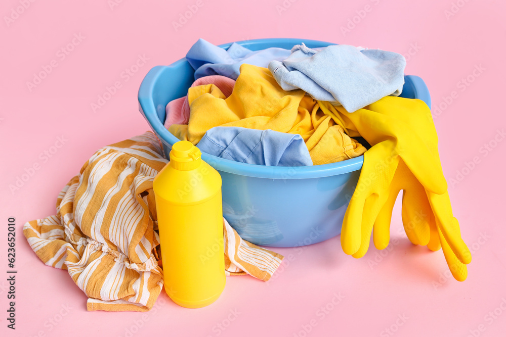 Plastic basin with dirty clothes, bottle of laundry detergent and yellow rubber gloves on pink backg