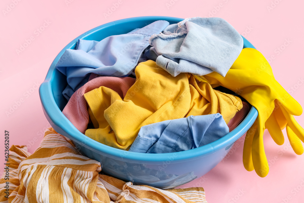 Plastic basin with dirty clothes and yellow rubber gloves on pink background