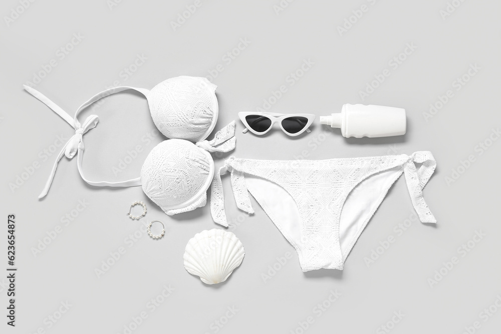 Stylish swimsuit, sunglasses, bottle of sunscreen cream, earrings and seashell on light background