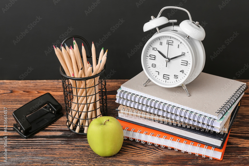 Different stationery, alarm clock and fresh apple on wooden table against black chalkboard