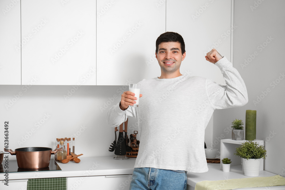 Young man with glass of milk showing muscles in kitchen