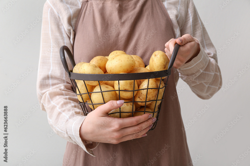 Woman holding basket with raw potatoes on light background