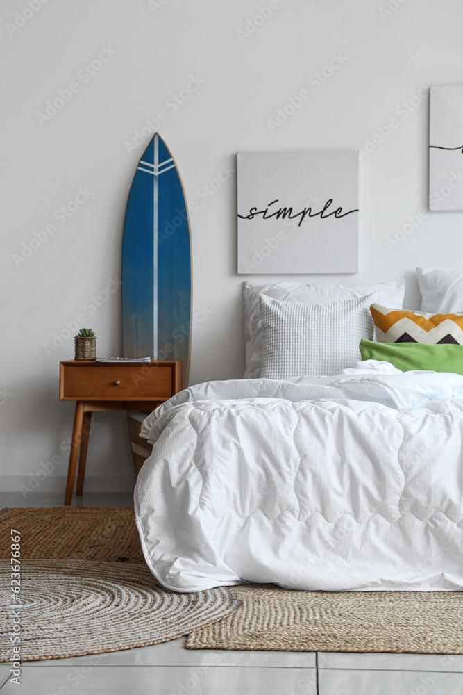 Cozy bed with white blanket and surfing board in interior of stylish bedroom