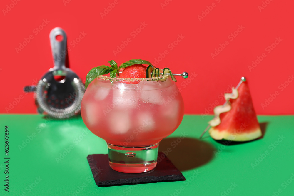 Glass of tasty watermelon cocktail with strainer on green table near red wall