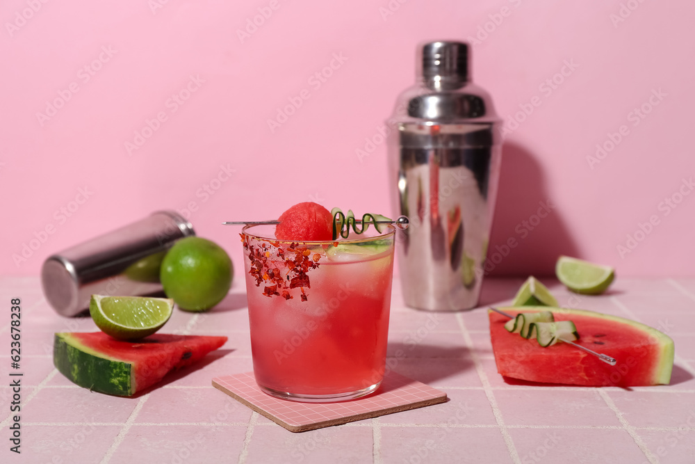 Glass of tasty watermelon cocktail with shakers and lime on tile table near pink wall