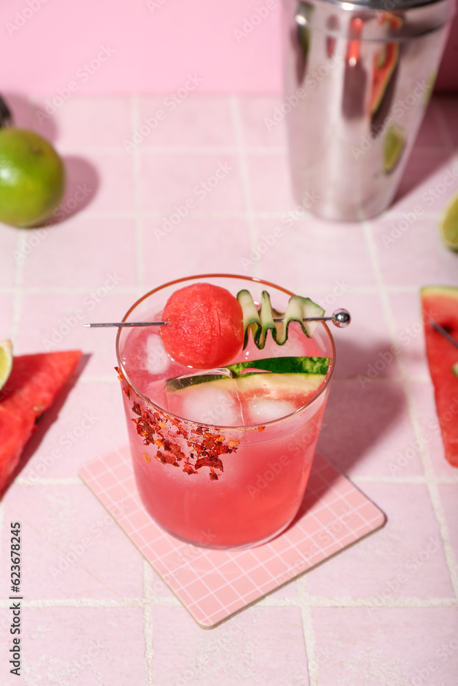 Glass of tasty watermelon cocktail with shaker and lime on pink tile background