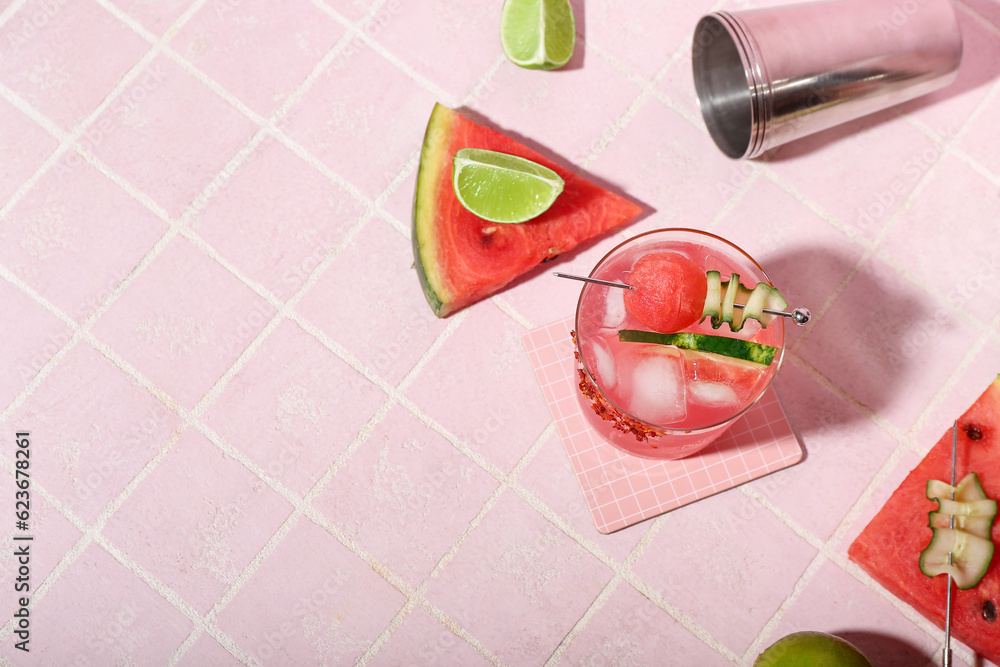 Glass of tasty watermelon cocktail with shaker and lime on pink tile background