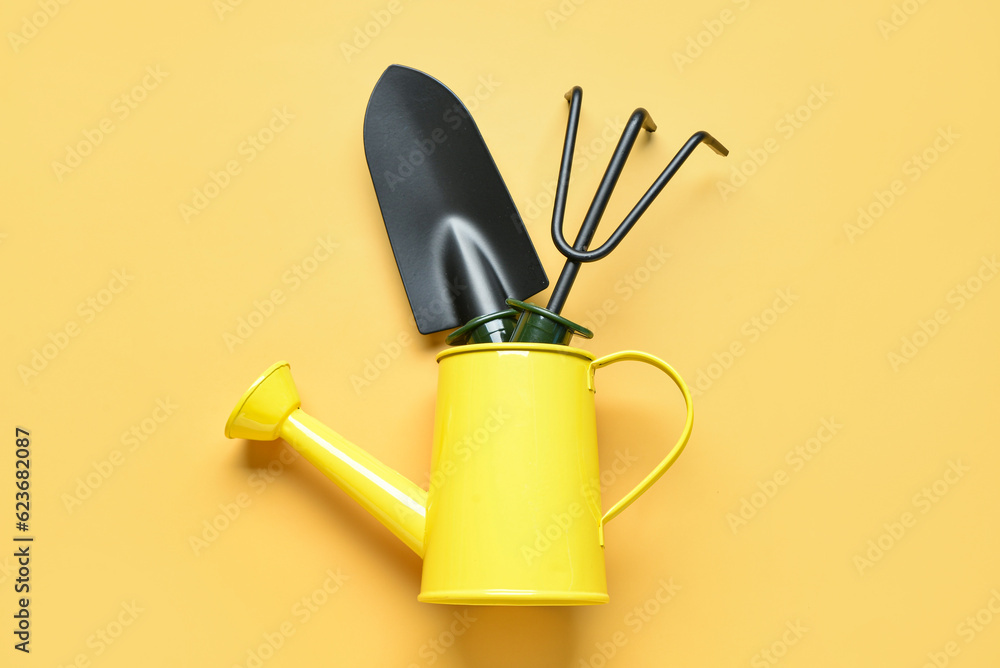 Watering can with gardening rake and shovel on yellow background