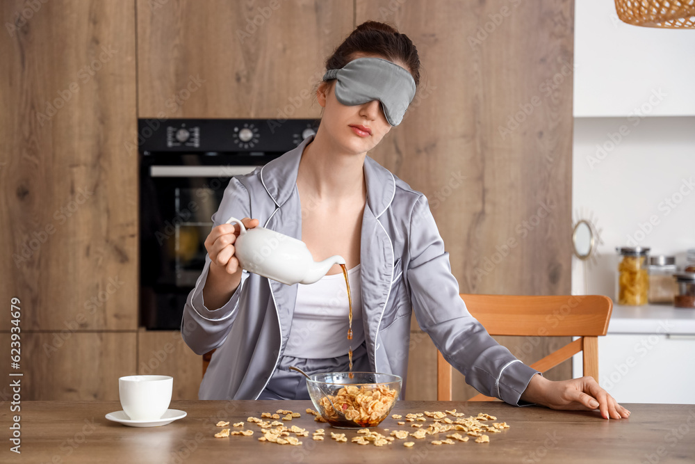 Young woman with sleeping mask pouring tea into bowl of cornflakes in kitchen