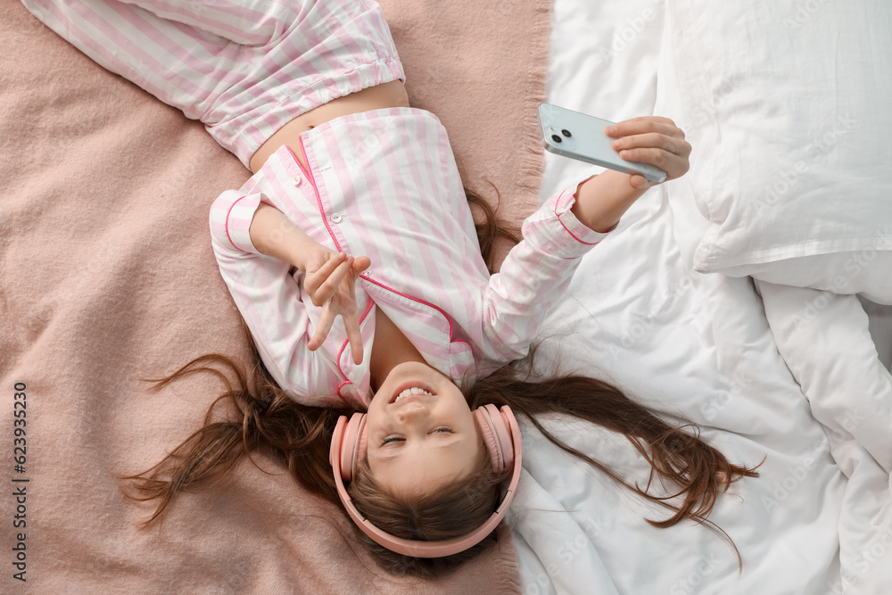 Little girl with headphones and mobile phone taking selfie on bed, top view