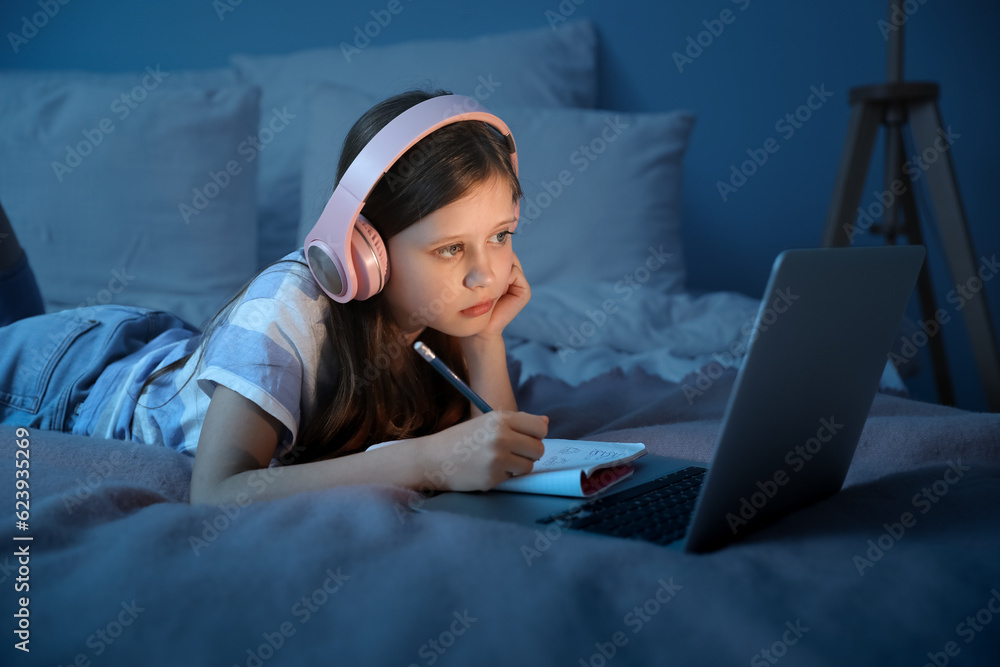 Little girl with headphones studying online in bedroom at night