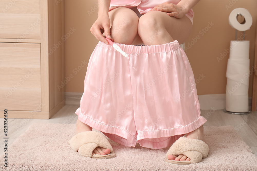 Young woman with pregnancy test sitting on toilet bowl in restroom