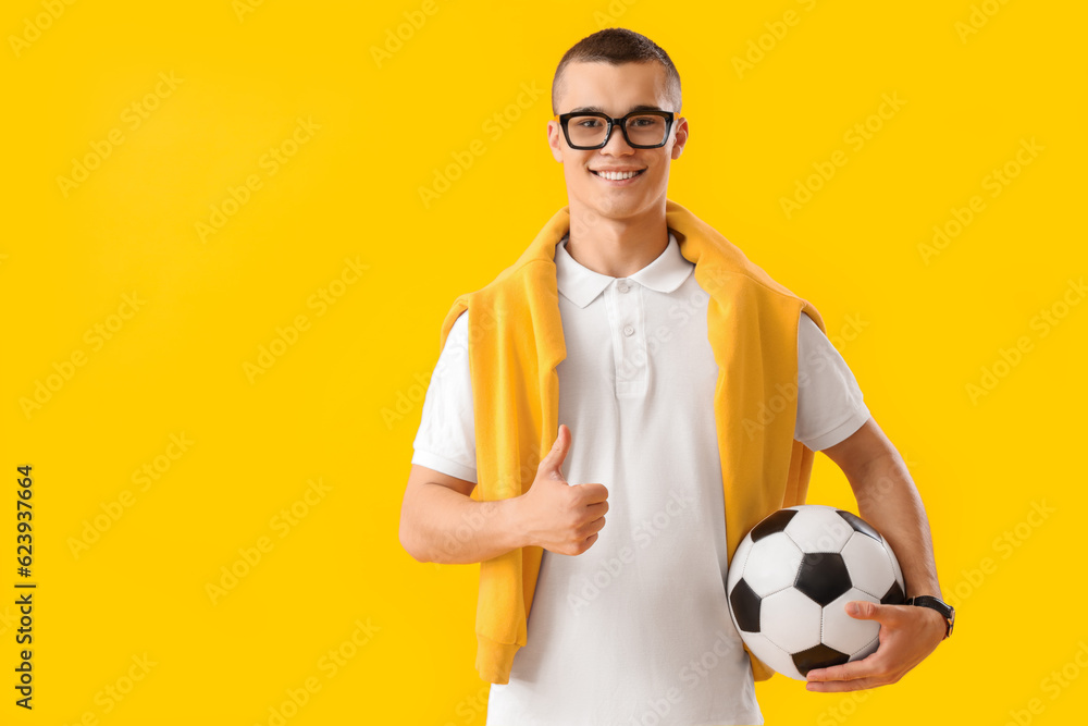 Male student with soccer ball showing thumb-up on yellow background