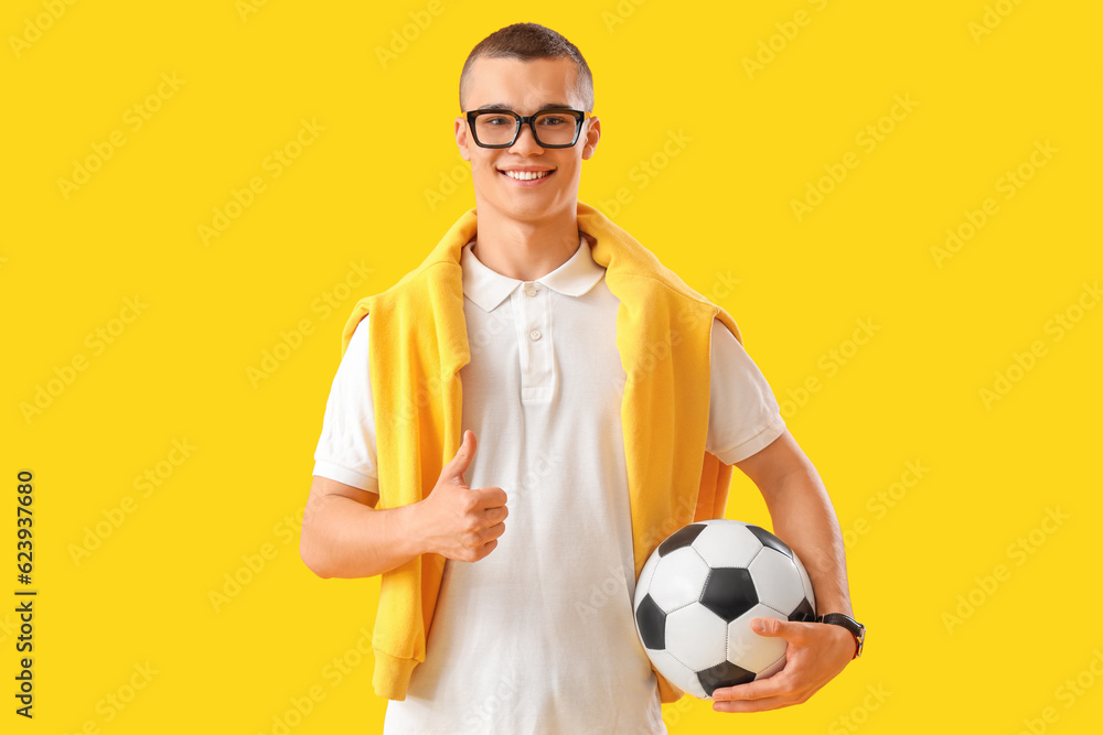 Male student with soccer ball showing thumb-up on yellow background