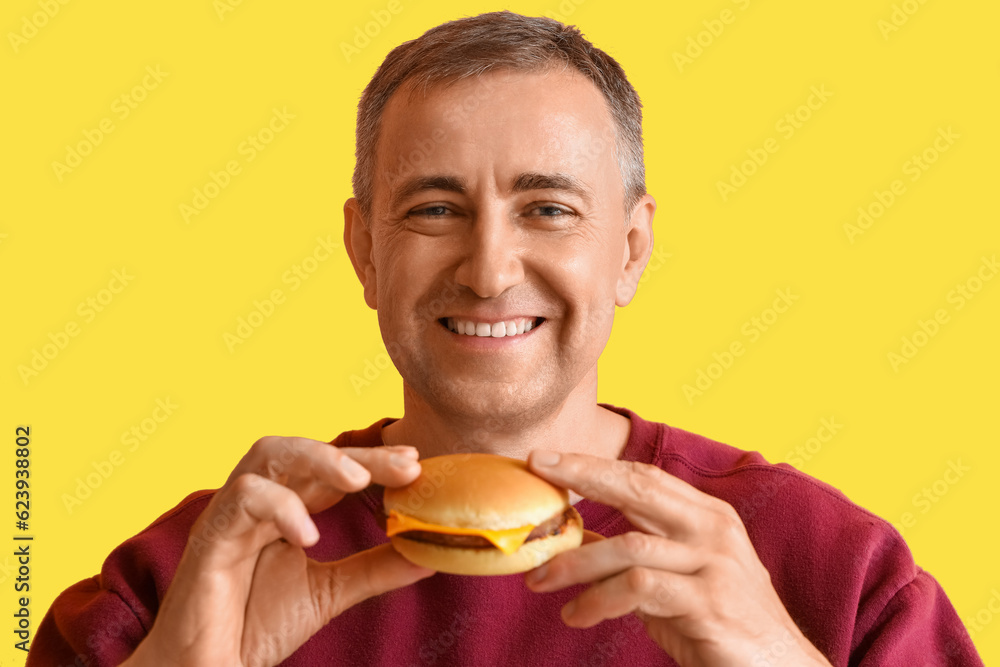 Mature man eating tasty burger on yellow background, closeup