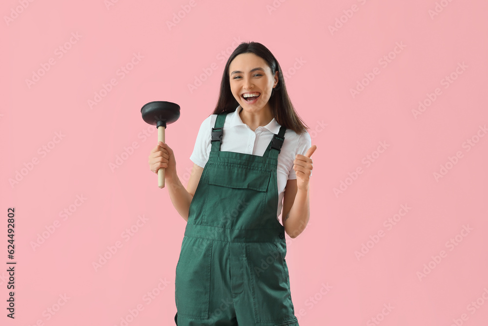 Happy female plumber with plunger showing thumb-up on pink background