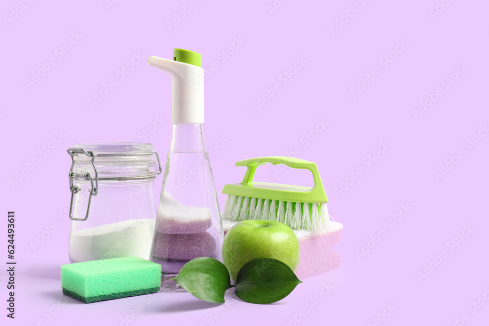 Bottle of vinegar, baking soda, sponges, brush and apple on lilac background