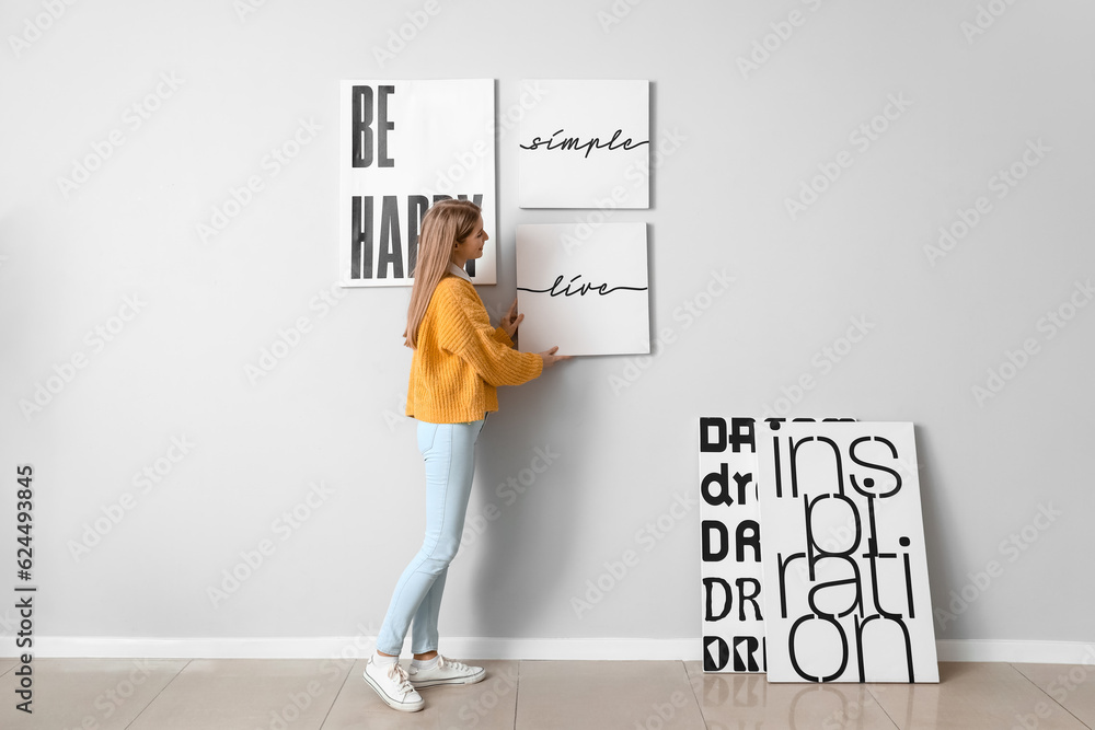 Young woman hanging poster on light wall at home