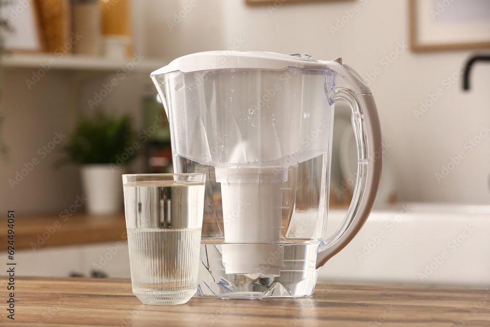 Glass of pure water and filter jug on wooden table in kitchen