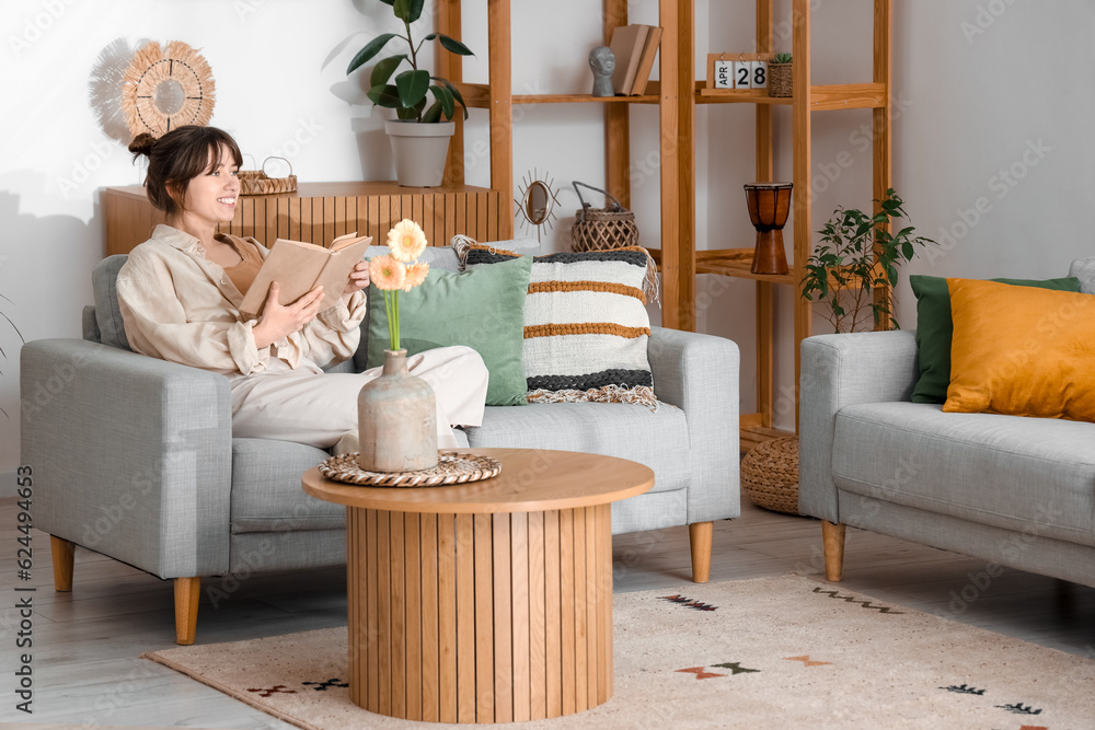 Happy young woman sitting on grey sofa and reading book in living room