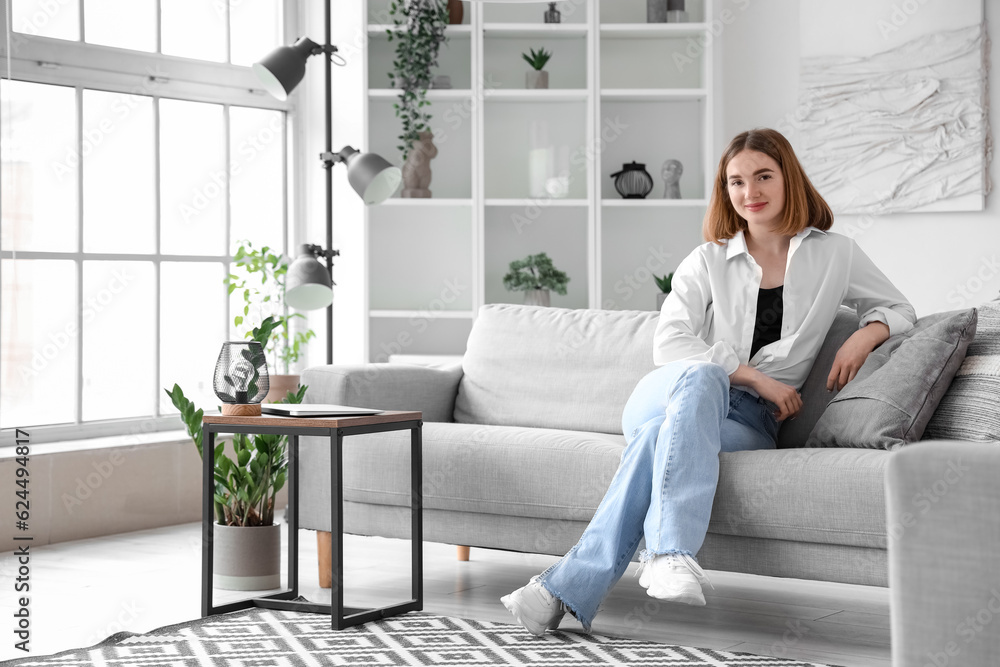 Pretty young woman sitting on grey sofa in interior of light living room