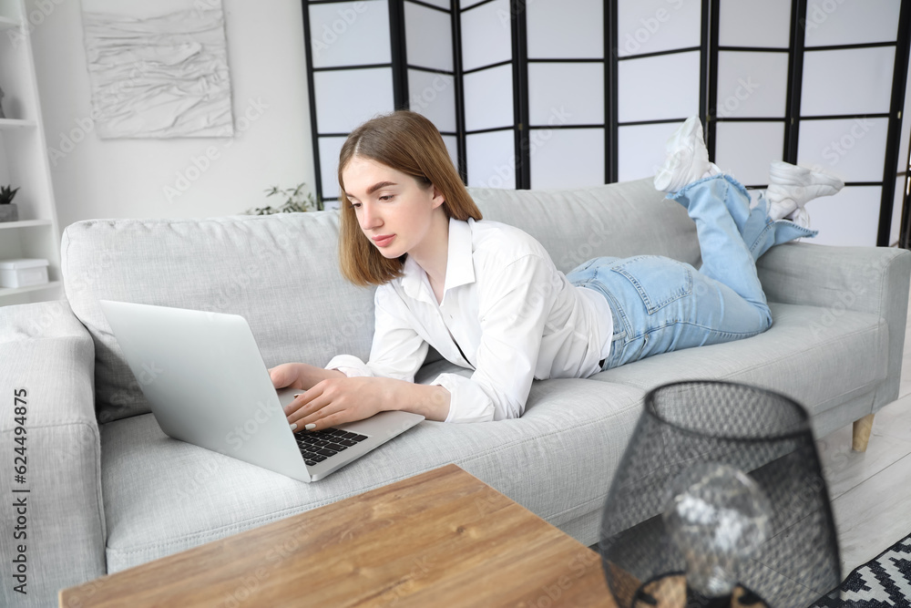 Pretty young woman lying on grey sofa and using modern laptop in light living room