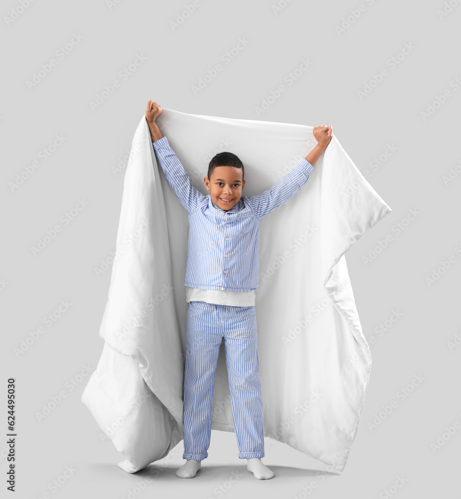 Little African-American boy in pajamas with soft blanket on light background