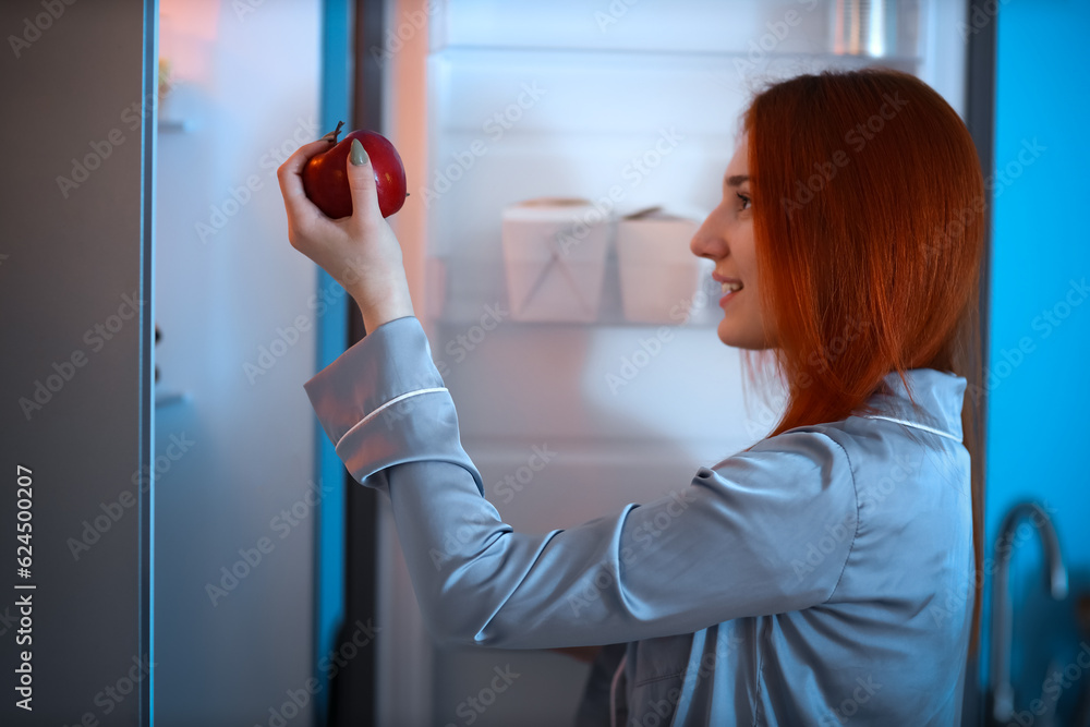 Young woman with apple near open fridge in kitchen at night