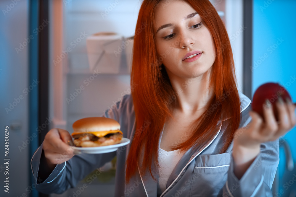 Young woman with burger and apple near open fridge in kitchen at night, closeup