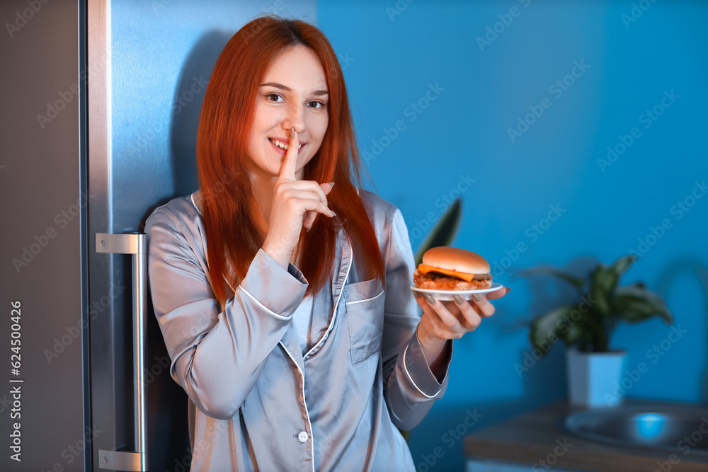 Young woman with tasty burger showing silence gesture near open fridge in kitchen at night