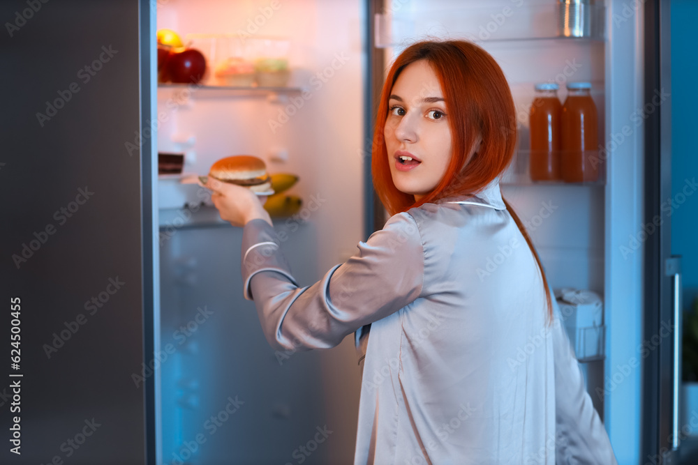 Young woman taking tasty burger from fridge in kitchen at night