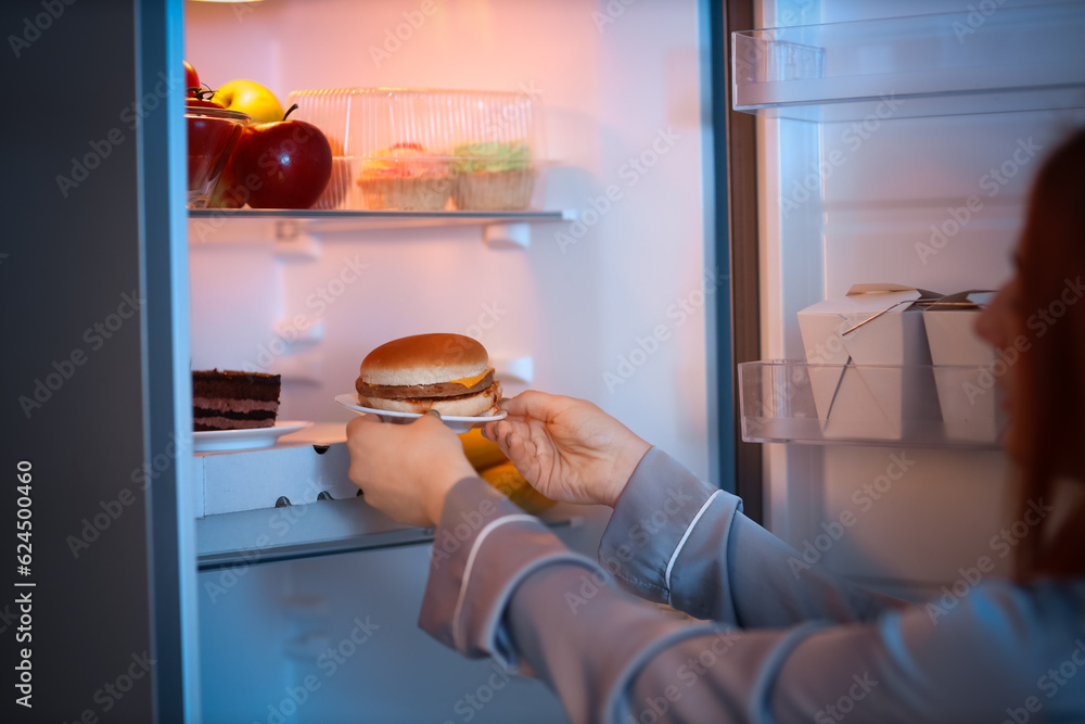 Young woman taking tasty burger from fridge in kitchen at night, closeup