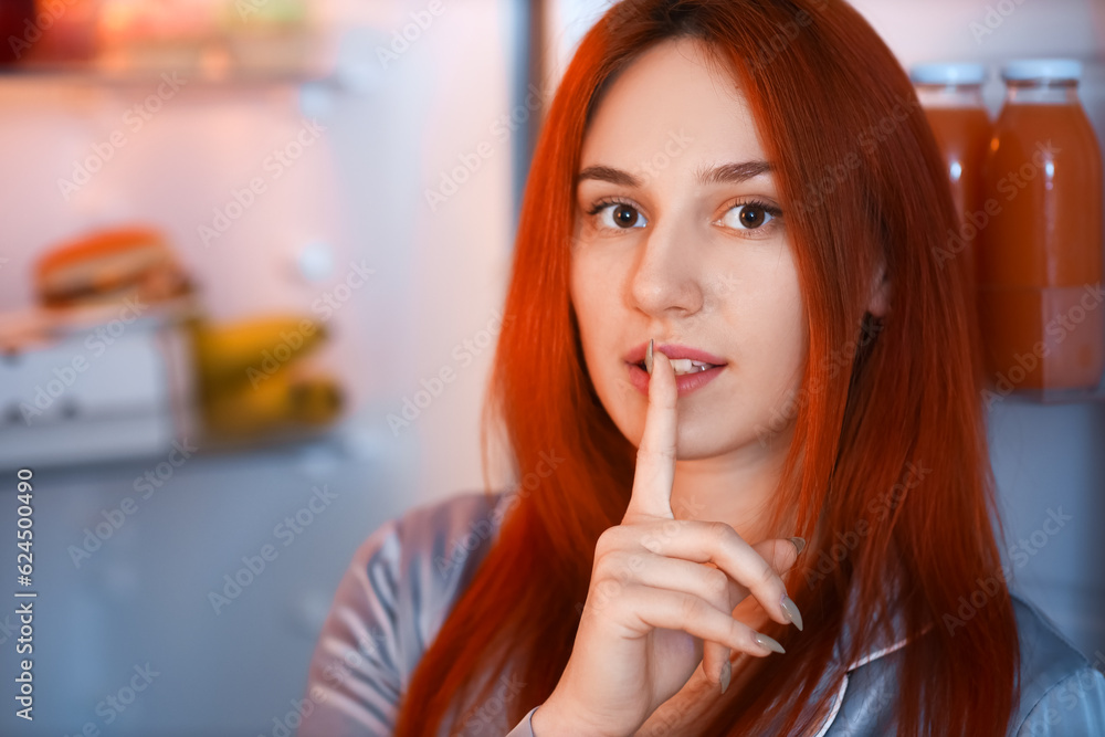 Young woman showing silence gesture near open fridge in kitchen at night, closeup