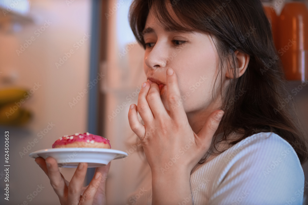 Young woman eating tasty doughnut near open fridge in kitchen at night, closeup