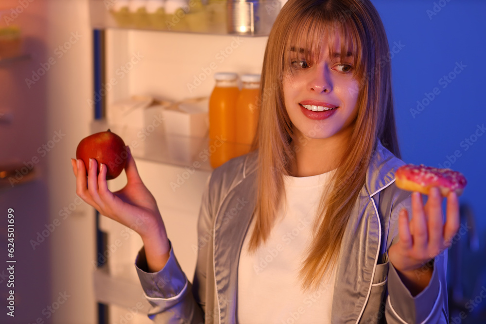 Young woman with tasty doughnut and apple near open fridge at night