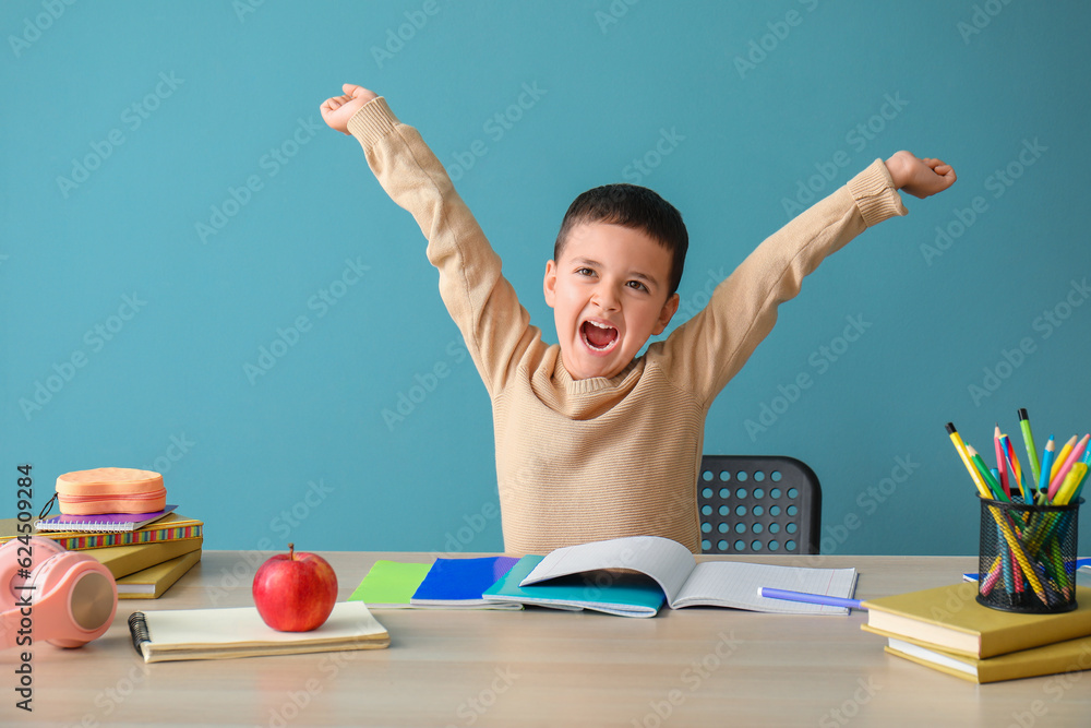 Happy little boy doing homework at table near blue wall