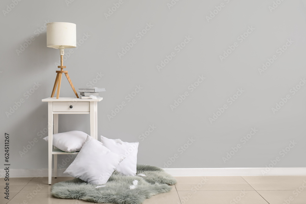 Bedside table with lamp and pillows near white wall