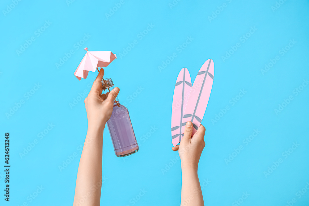 Female hands with bottle of cocktail and mini surfboards on blue background