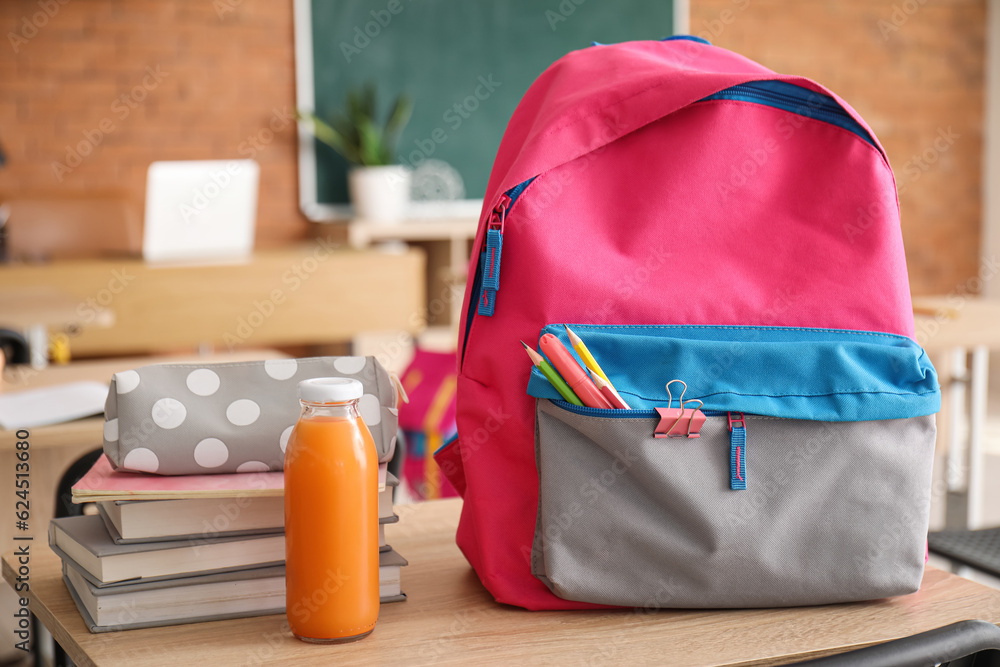 Backpack with bottle of juice and stationery on desk in classroom