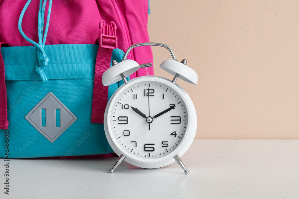 Colorful school backpack with alarm clock on white table near orange wall