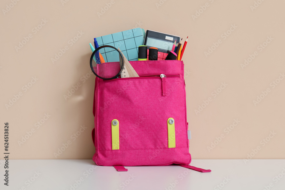 Pink school backpack with different stationery and magnifier on white table near orange wall