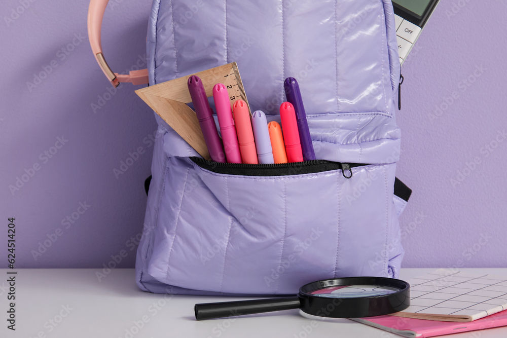 School backpack with different stationery and headphones on white table near purple wall