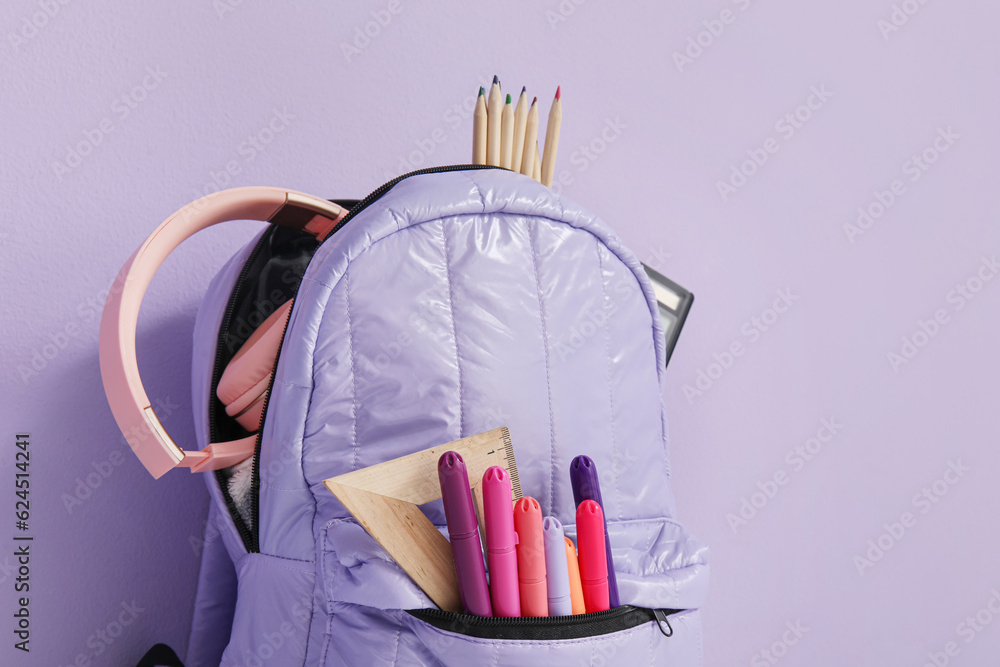 School backpack with different stationery and headphones on purple background